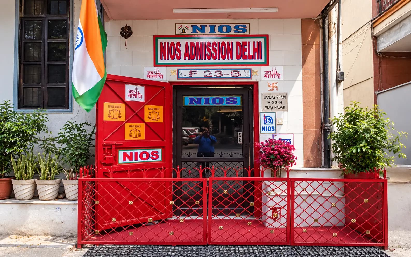 Office entrance with a red door and gate below a sign reading NIOS Admission Delhi, F 23-B